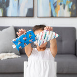 A child holds up Bumkins Reusable Snack Bag 4 Pack: Disney Mickey & Friends Crew, covering their face while sitting in front of a couch.