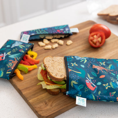 Bumkins Reusable Snack Bag 4 Pack: Jungle and Animal Print holds veggies, snacks, & a sandwich on a wood cutting board on a kitchen counter.