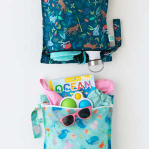 Bumkins Wet Bag 2 Pack: Ocean Life and Jungle holds toys, swimsuits, sunglasses, books, a towel, and a water bottle on a white background.