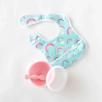 A blue Bumkins Starter Bib 2 Pack: Rainbows & Unicorns is paired with a pink baby bowl and spoon set on a white background.