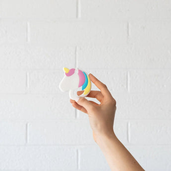 A hand holds a Bumkins Silicone Teether: Unicorn against a white brick wall.