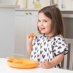 A girl in a Bumkins Junior Bib: Hearts sits at a white table, eating from an orange plate with orange utensils.