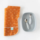 A folded orange Bumkins Junior Bib 2 Pack: Desert Boho lays next to a gray plate and utensils on a white background.