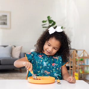 A young girl with curly hair and a bow wearing a Bumkins Junior Bib: Jungle eats from an orange plate using a fork at a white table.