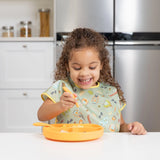 A child smiles while using a fork to eat from an orange plate, wearing a Bumkins Junior Bib: Camp Gear at a white table.