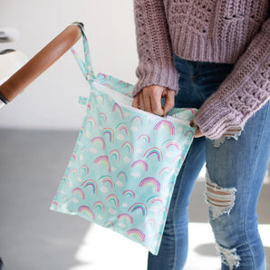 A person in a pink sweater and ripped jeans unzips a Bumkins Wet Bag 2 Pack: Hearts and Rainbows hanging from a stroller handle.